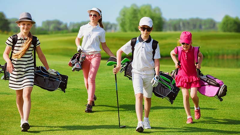 kids,walking,on,fairway,with,bags,at,golf,school